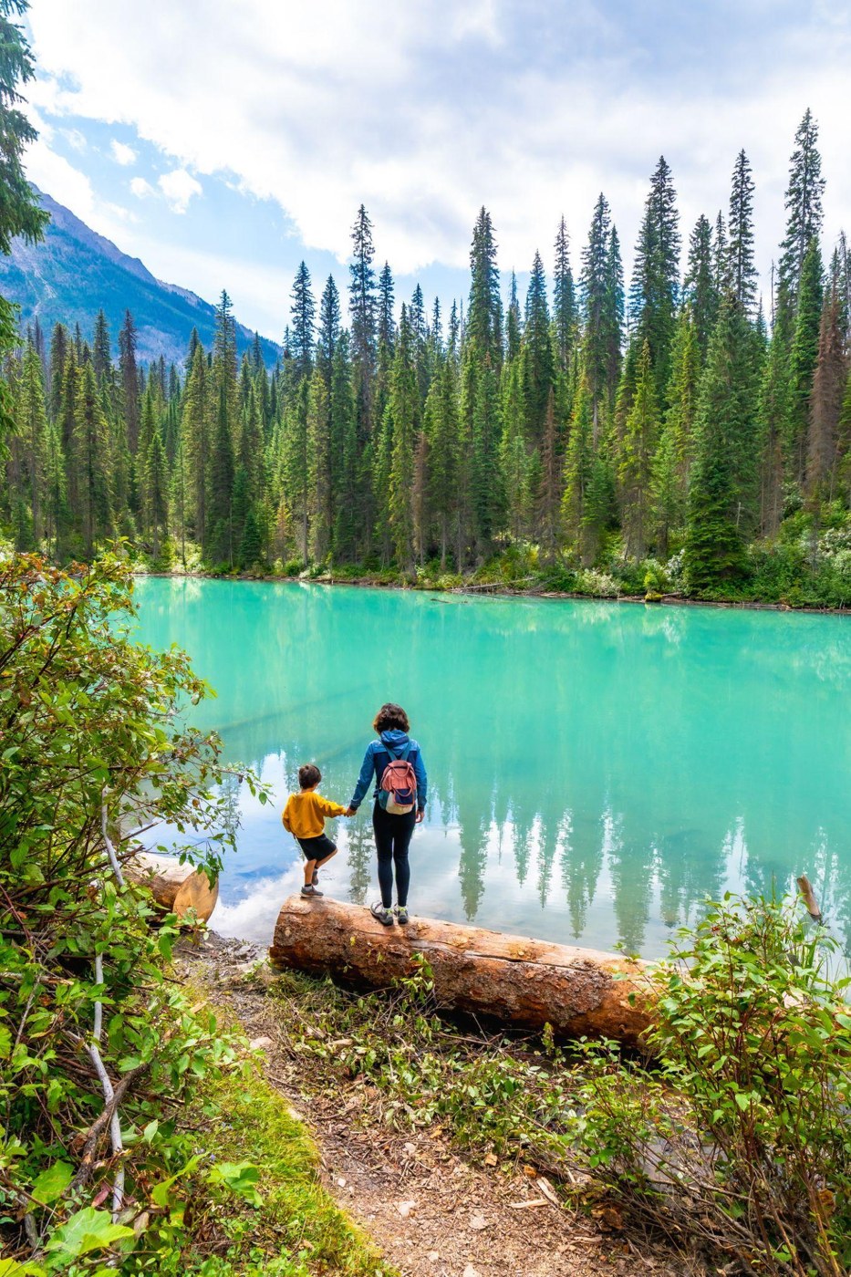 Two people beside a turquoise lake surrounded by evergreen forest and mountains